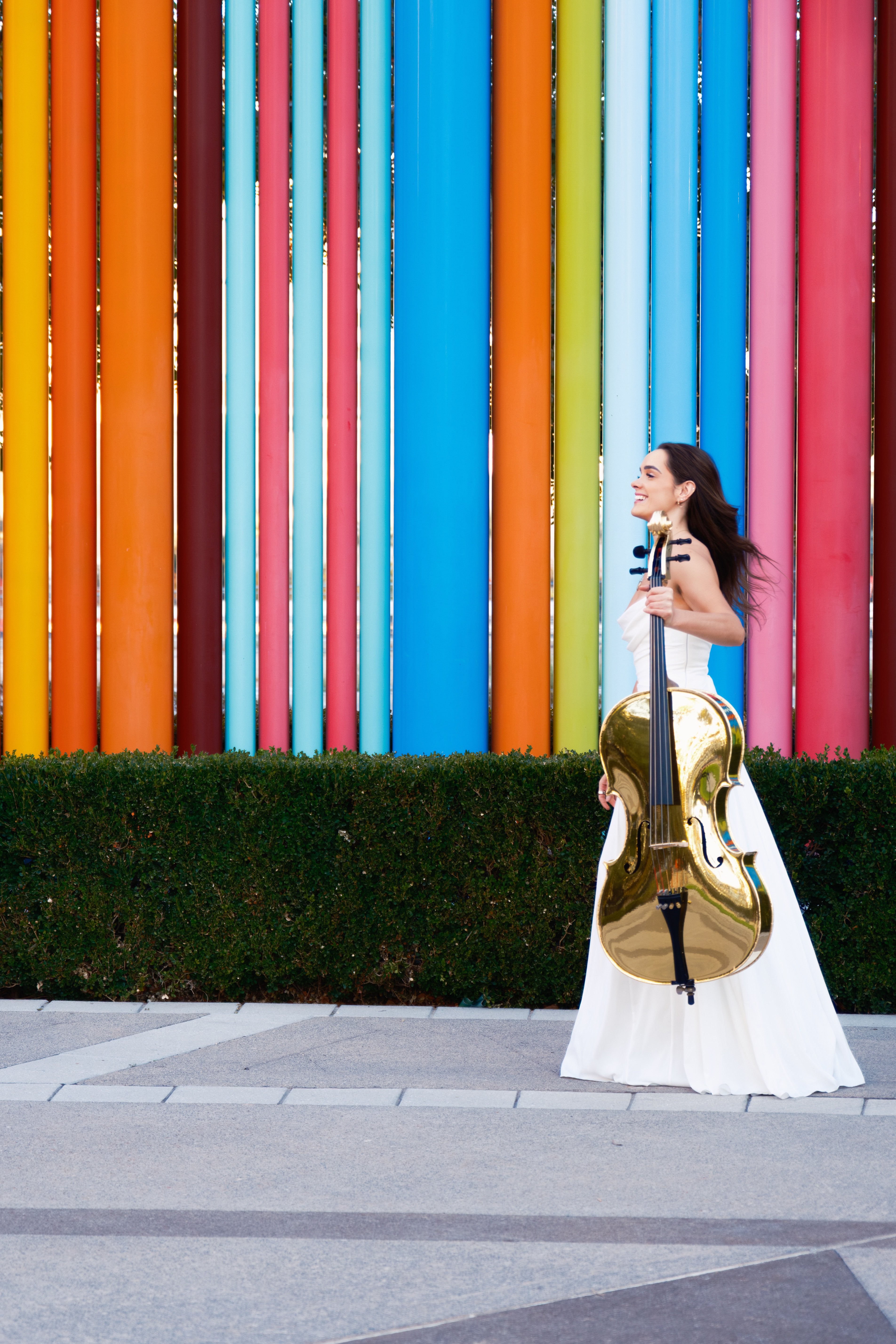 Golden Cello Girl in a white gown holding her signature gold cello in front of a colorful sculpture in Los Angeles