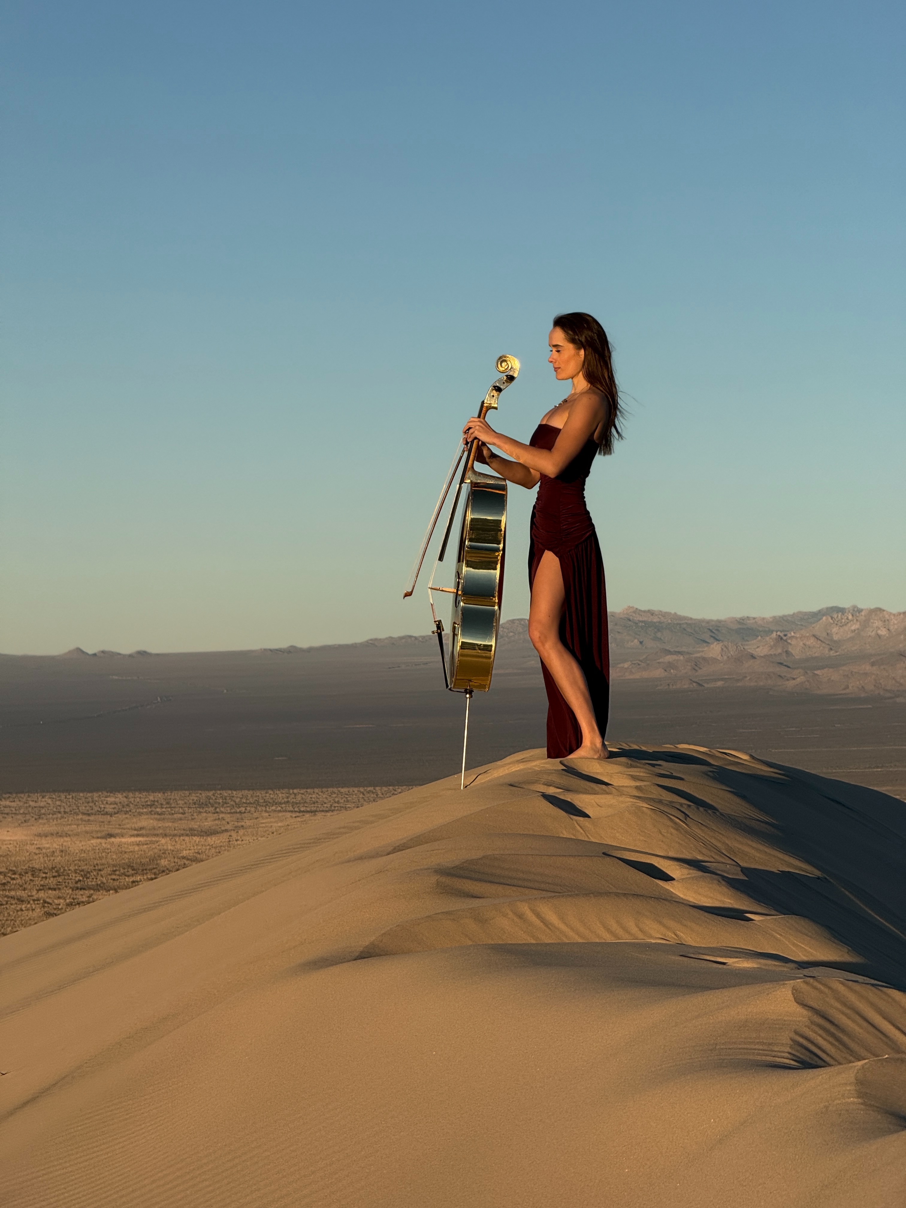 Cellist standing atop sand dune in burgundy dress with golden cello against blue sky